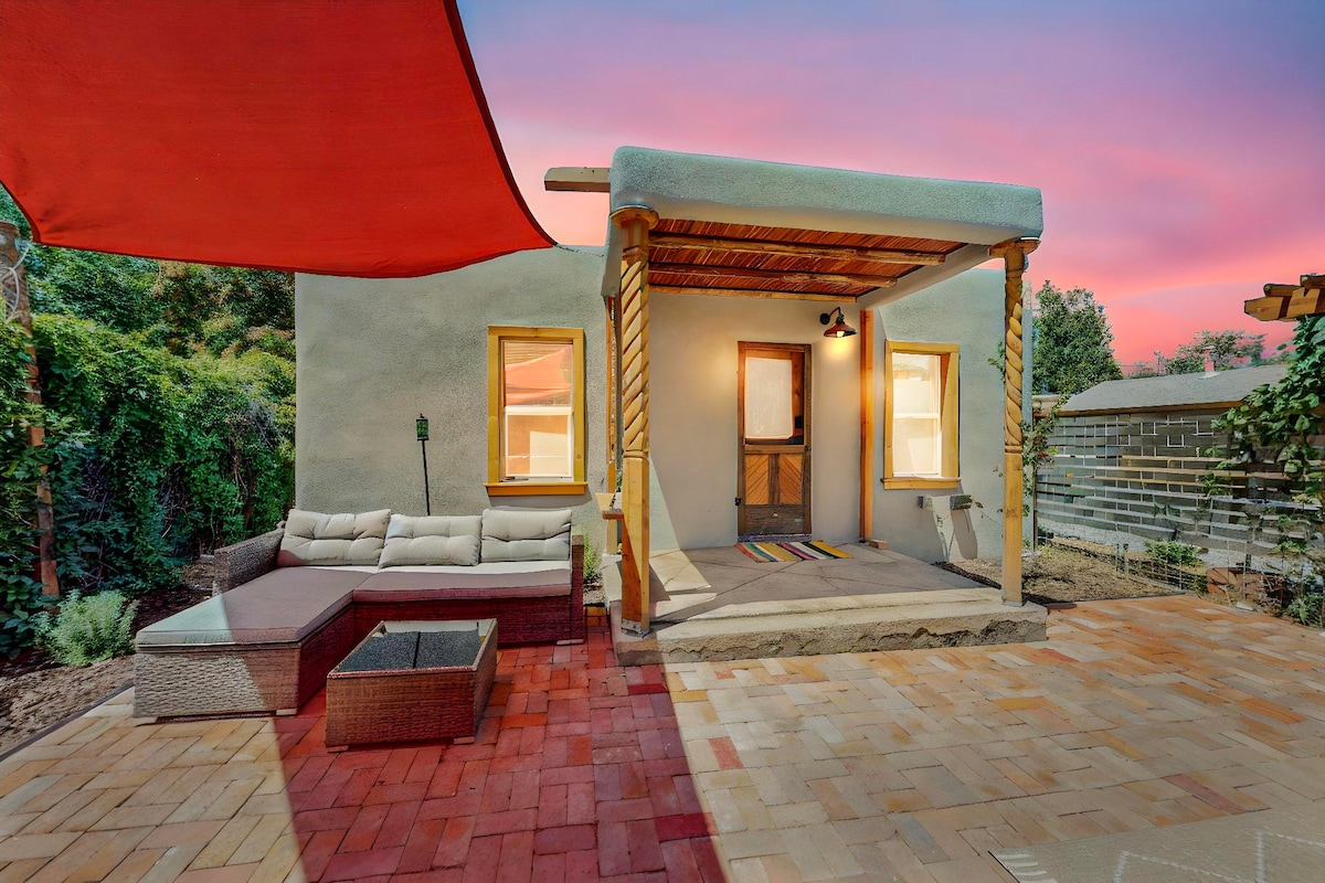A cozy outdoor seating area features a sectional sofa and a fire pit, set against a backdrop of lush greenery. The building's rustic façade is complemented by colorful accents and soft lighting, creating a welcoming entrance under a vibrant sunset sky.