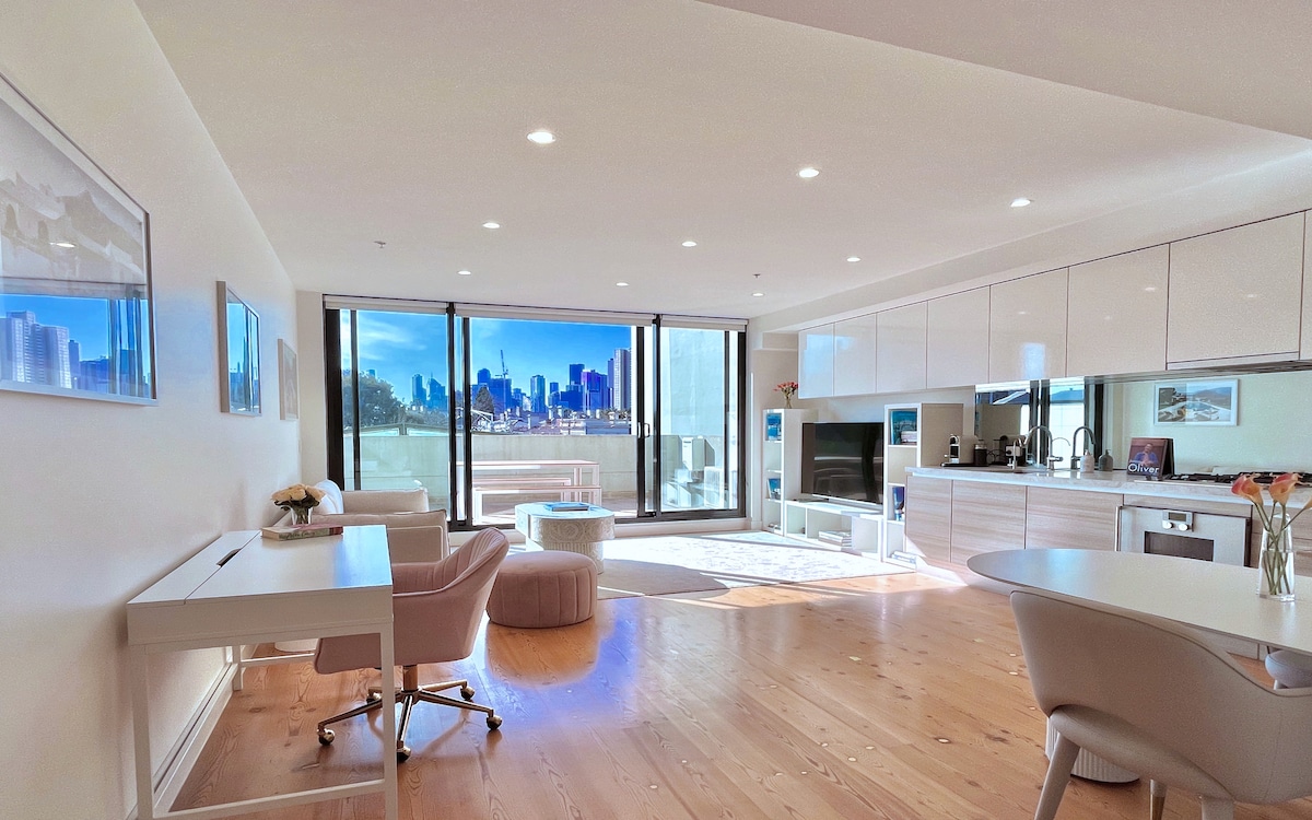An open-plan living area features large glass doors that provide access to an outdoor terrace and stunning views of the Melbourne skyline. Light wood flooring complements modern furnishings, including a desk and dining area, while natural illumination enhances the space.