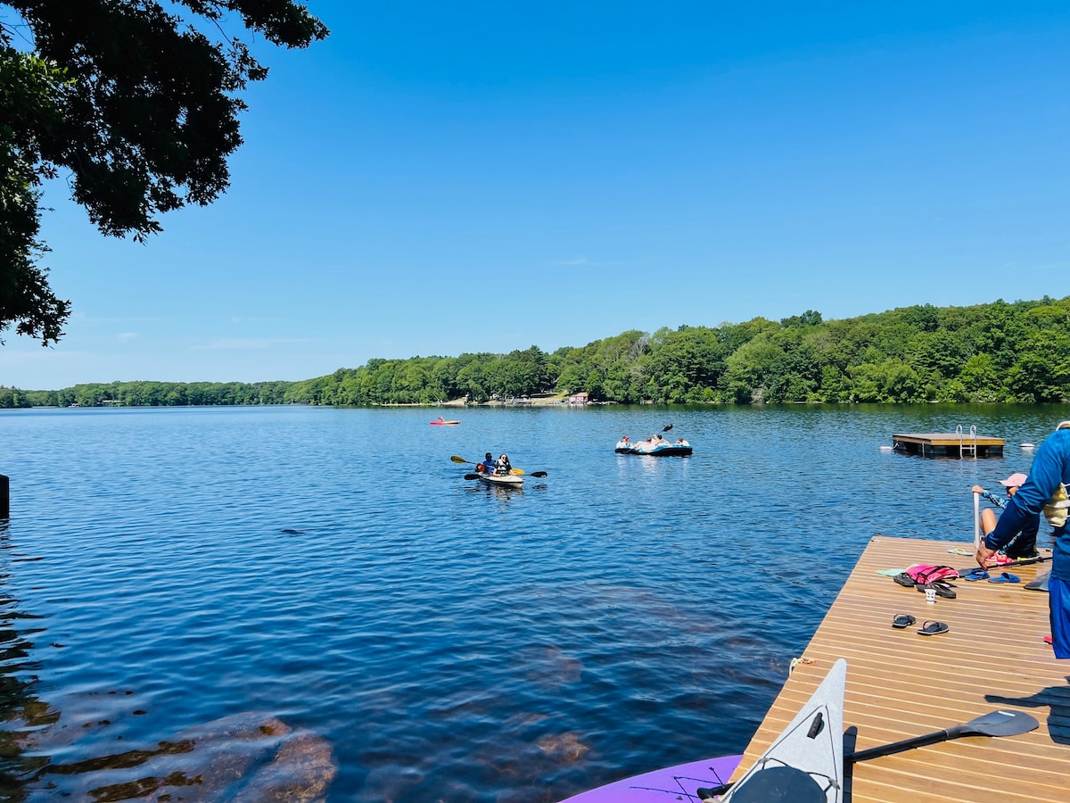 A serene lake scene is depicted with calm waters reflecting the blue sky. Kayakers navigate the lake, surrounded by lush green trees. A wooden dock juts out, providing a vantage point for enjoying the tranquil environment and activities on the water.