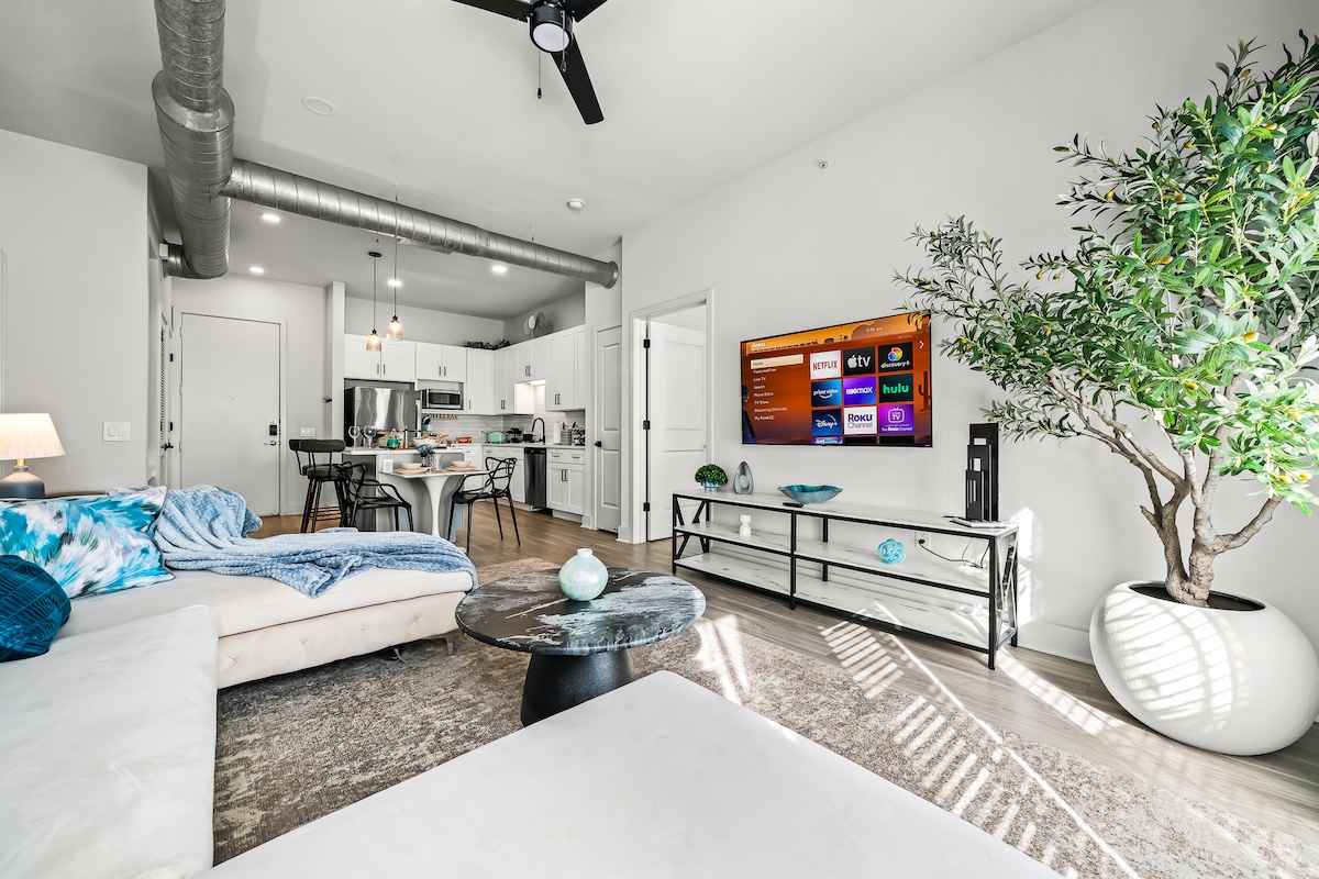 A bright living area showcases a spacious sectional sofa with decorative pillows, centered around a sleek coffee table. A large smart TV is mounted on the wall, while a dining area with modern seating is visible in the background, illuminated by natural light from large windows.