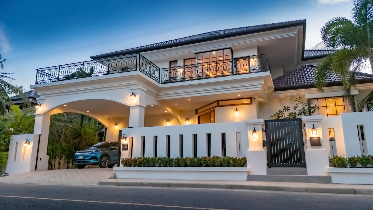 The villa's elegant exterior is characterized by a two-story structure with a tiled roof and large windows. A spacious driveway leads to a covered parking area. Lush greenery surrounds the entrance, enhancing the welcoming appearance of the home.