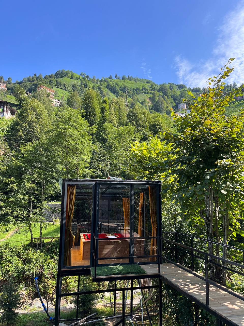 A modern glass structure is positioned on a raised platform, surrounded by lush greenery. The vibrant blue sky and distant hills enhance the serene natural environment, while the transparent walls provide unobstructed views of the surrounding landscape.