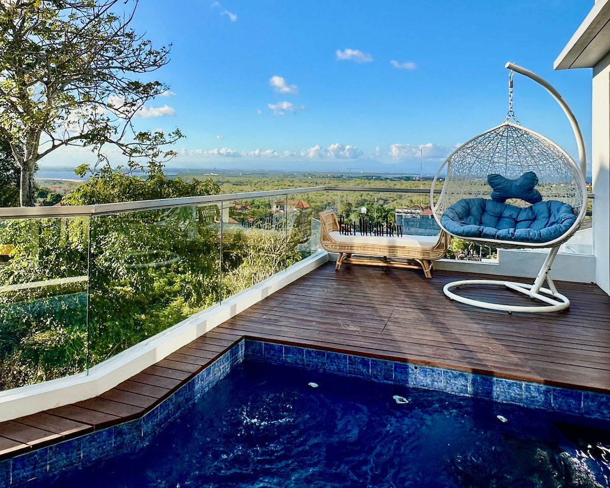 An inviting outdoor space features a hanging chair and a sun lounger, overlooking scenic views of greenery and distant hills. The private pool is surrounded by a wooden deck, offering a serene spot for relaxation under the blue sky.