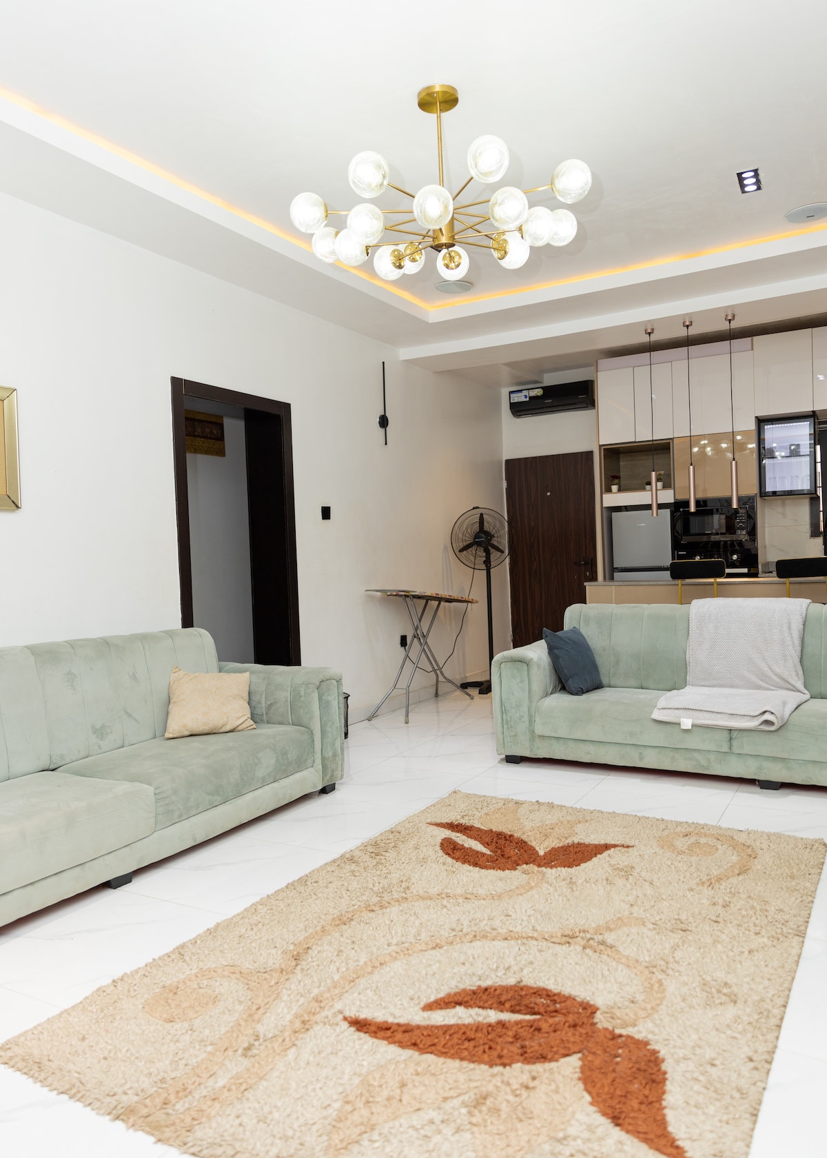 A bright living area features two soft green sofas set on a neutral-toned rug with a floral pattern. A modern chandelier provides ambient lighting, and a small dining table is visible in the background, alongside an electric fan and sleek kitchen appliances.