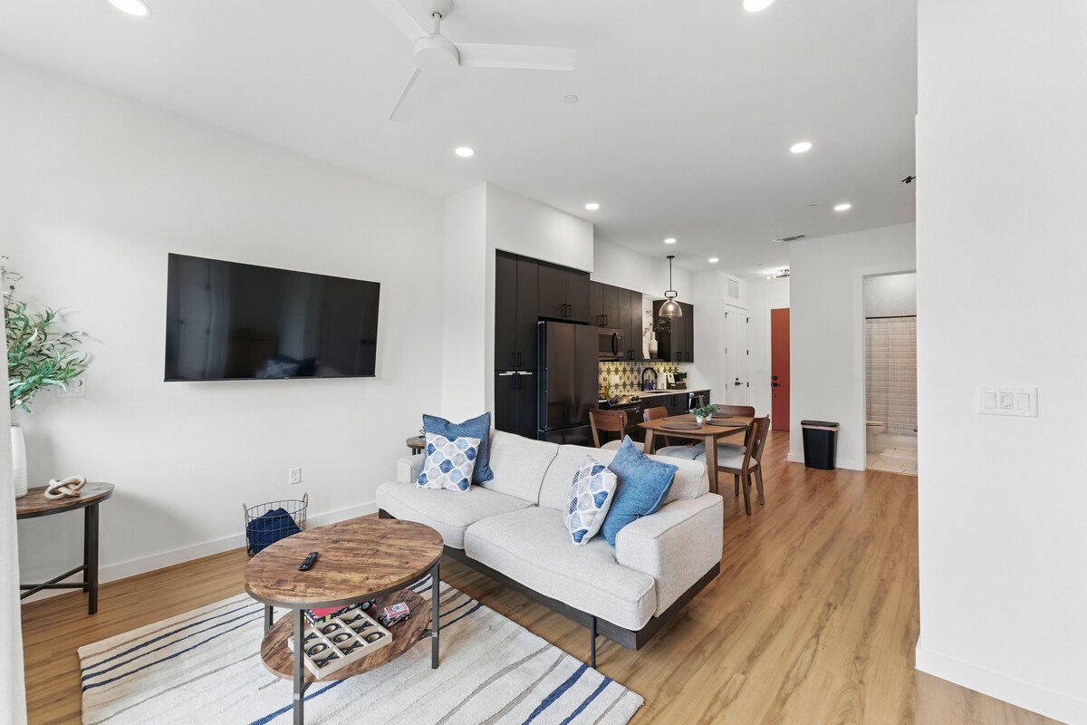 A contemporary living area is featured, showcasing a light-colored couch adorned with blue and white accent pillows. A wooden coffee table sits in front, accompanied by a mounted flat-screen TV on the wall. The open layout extends towards a dining area and kitchen in the background.