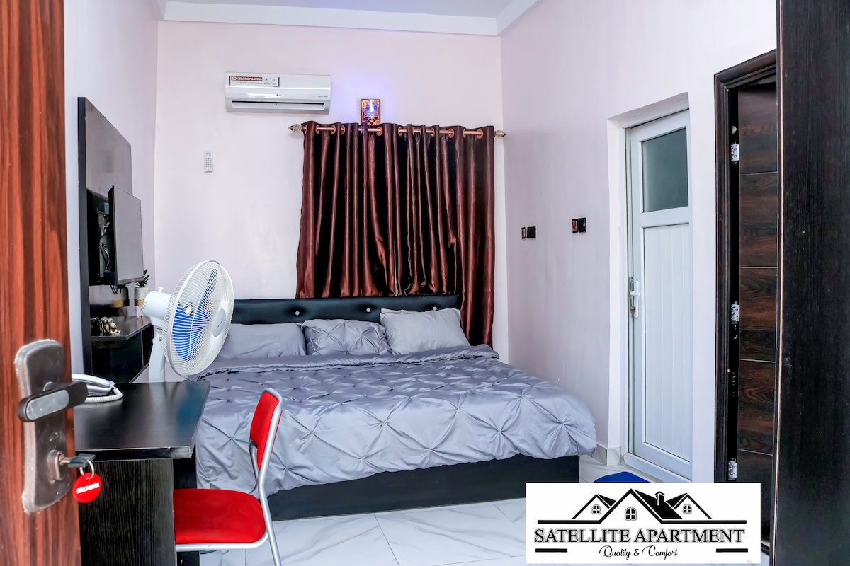 A cozy sleeping area features a spacious bed with grey linens, complemented by dark curtains that filter natural light. A small desk and chair are positioned adjacent to the bed, while a wall-mounted television provides entertainment. Air conditioning is visible above for climate control.