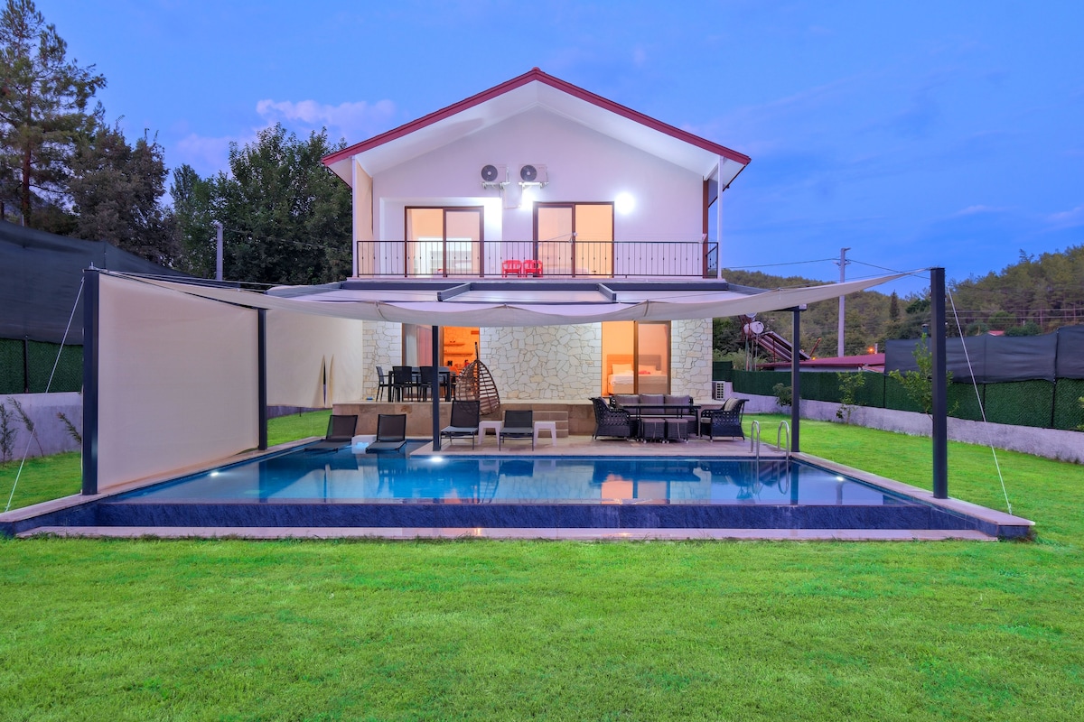 A spacious villa is showcased with an inviting pool surrounded by a well-maintained lawn. The exterior features large windows with a balcony above, complemented by shaded seating areas beside the pool. Lush greenery enhances the relaxing outdoor setting.