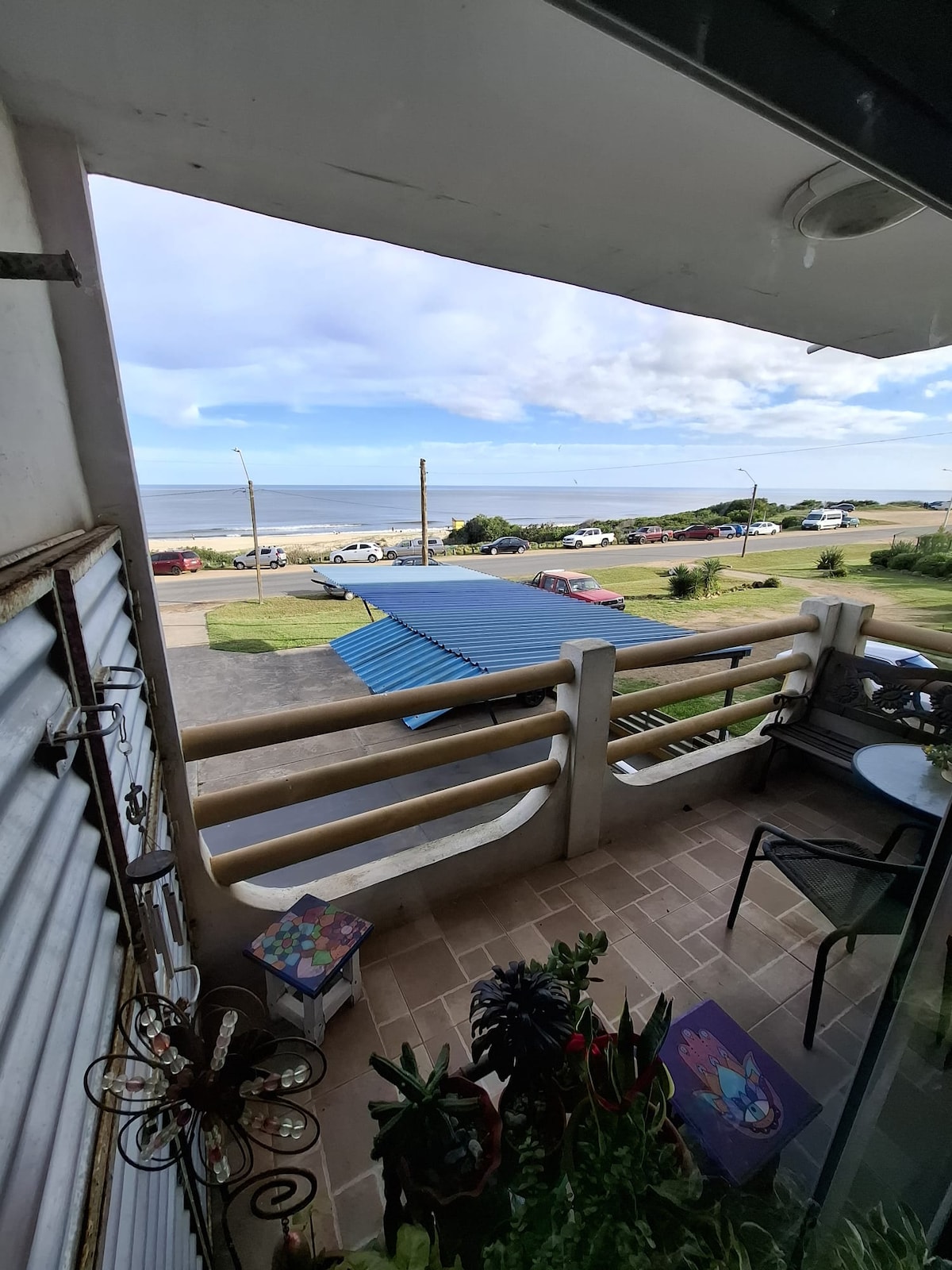 A balcony view features outdoor seating with a small table and potted plants. The scene overlooks a grassy area and the coastline, with vehicles visible along the beach road. The sky is partially cloudy, complementing the tranquil atmosphere.