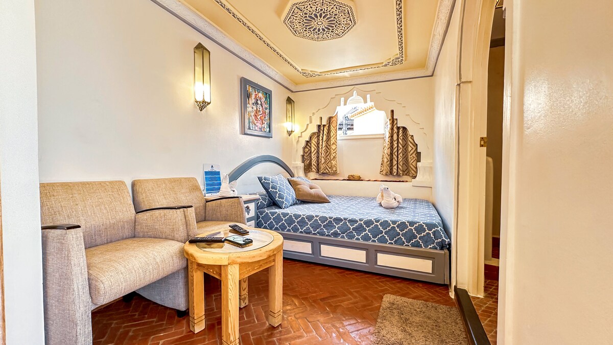 A spacious guest room features a large bed with patterned bedding, complemented by decorative wall carvings. A cozy seating area consists of two armchairs and a round table. Natural light filters in through a window with traditional curtains, enhancing the serene atmosphere.