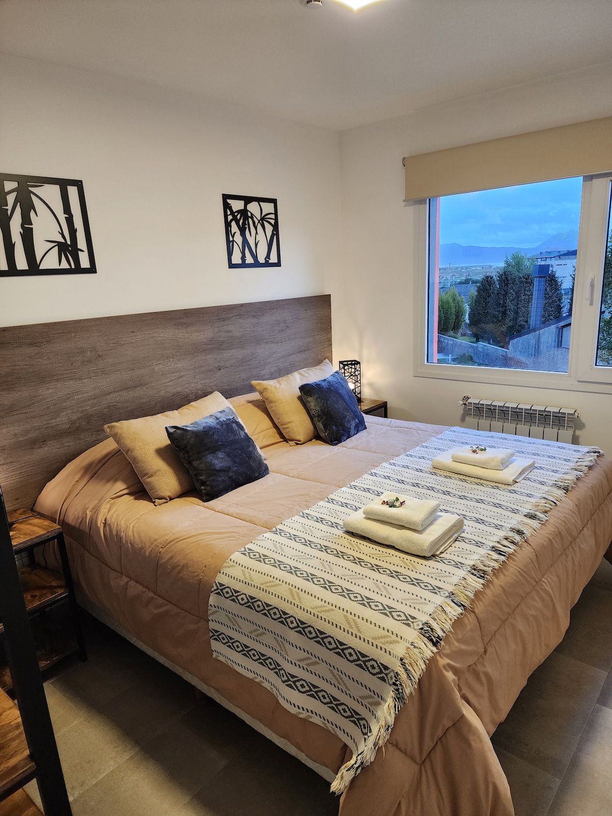 A cozy bedroom features a spacious bed with beige and blue accent pillows. A woven throw blanket rests across the foot of the bed. Natural light filters through a window with a scenic view, complemented by decorative wall art of bamboo.