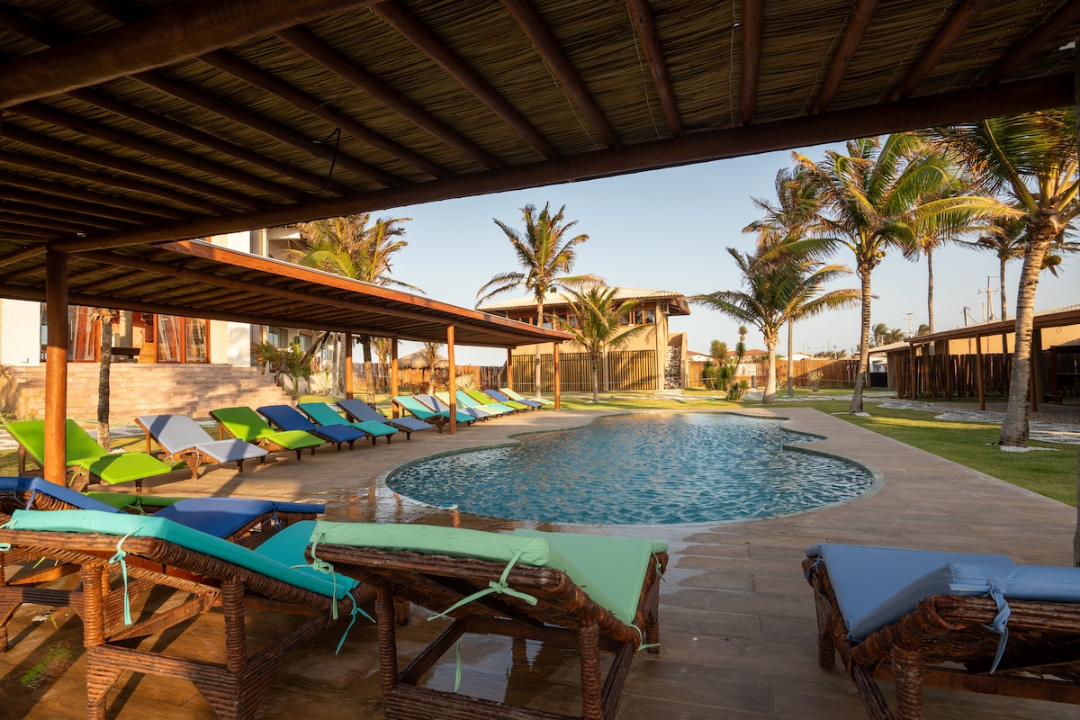 A sunny outdoor area features a curvilinear pool surrounded by colorful loungers in shades of blue and green. Palm trees provide gentle shade, and the wooden pergola adds architectural interest. The spacious layout invites relaxation and enjoyment in the coastal environment.