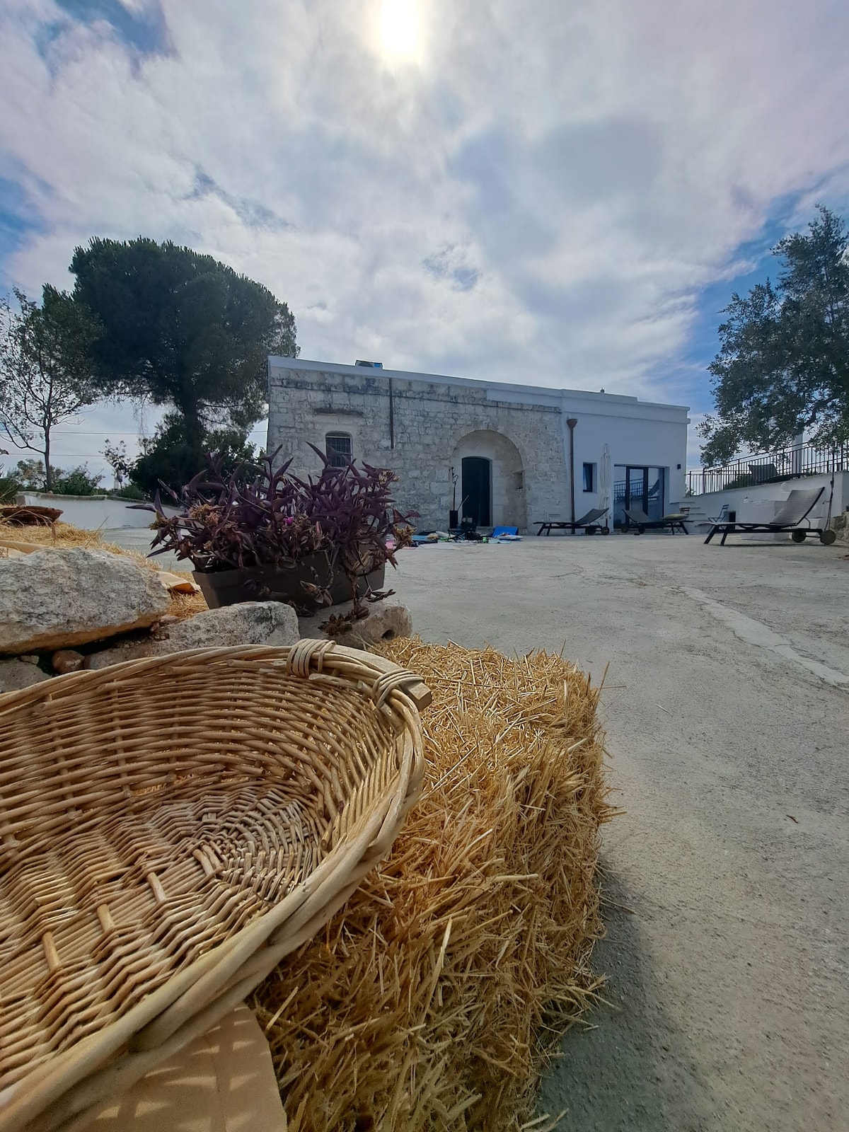 A rustic stone building is viewed from a distance, framed by a cloud-filled sky. Straw bales and a woven basket are placed in the foreground, adding to the outdoor setting. Wooden furniture is visible, suggesting a spacious area for relaxation.