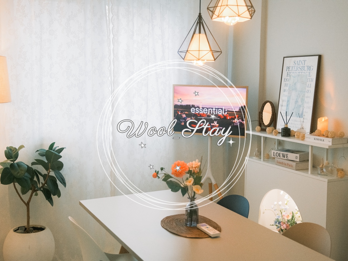A bright interior space features a white table set for dining and adorned with flowers. A television screen displays vibrant imagery, while natural light filters through sheer curtains. Decorative lights remind of a cozy ambiance, complemented by a stylish plant nearby.