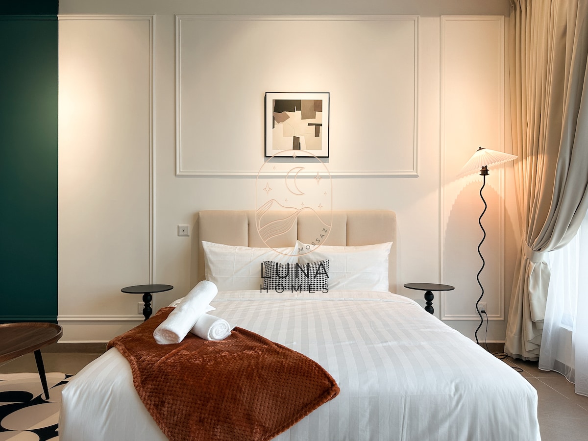 A spacious bedroom features a queen bed with a soft quilt and two rolled towels. A minimalistic art piece hangs above the bed. Nightstands on either side are paired with stylish lamps, and natural light fills the room from a nearby window with sheer curtains.