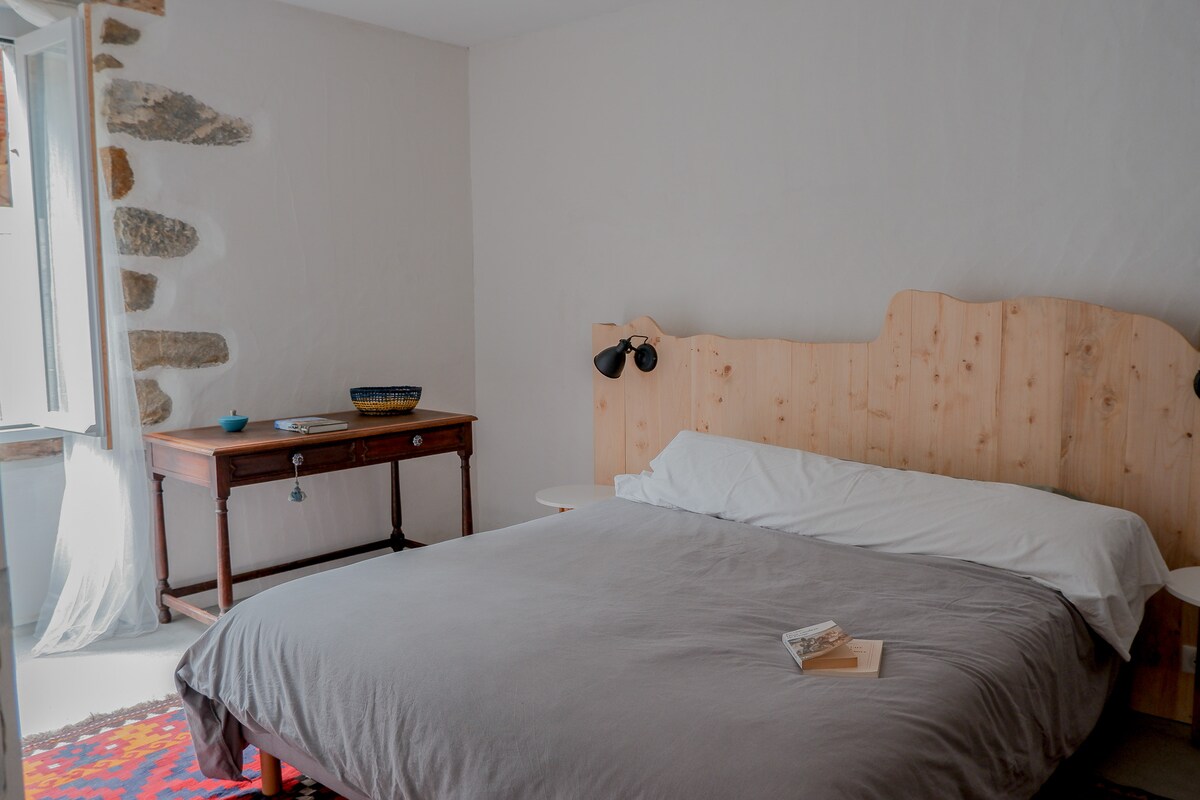 A spacious bedroom features a wooden headboard and a comfortable bed dressed in soft linens. Natural light flows in through a window adorned with sheer curtains, illuminating a wooden table placed beside the bed, enhancing the serene ambiance.