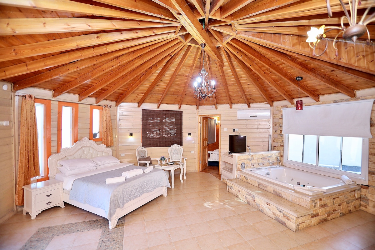 A spacious studio room features a circular wooden ceiling with exposed beams. A comfortable bed with light linens and decorative pillows is positioned next to a wooden nightstand. A jacuzzi and a seating area are included, with natural light filtering through large windows.