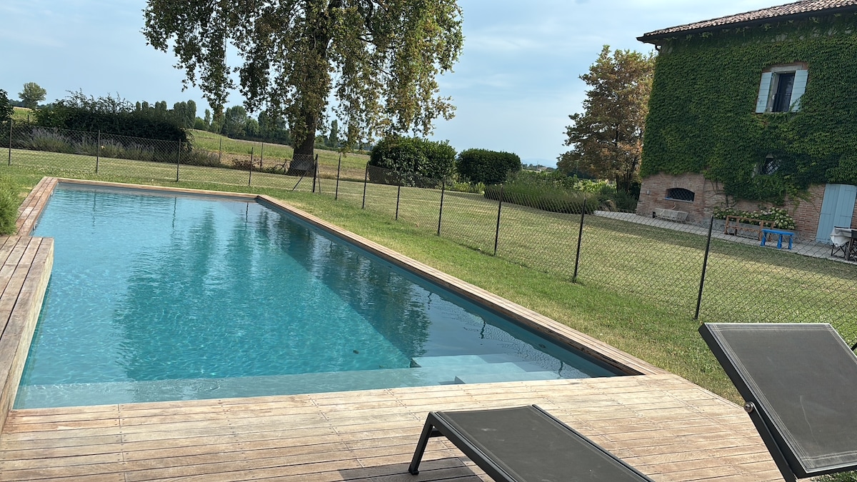 A serene outdoor space features a rectangular swimming pool, surrounded by a wooden deck and a grassy area. The pool reflects the sky, with a fence providing a boundary. Lush trees and greenery are visible in the background, adding to the tranquil setting.