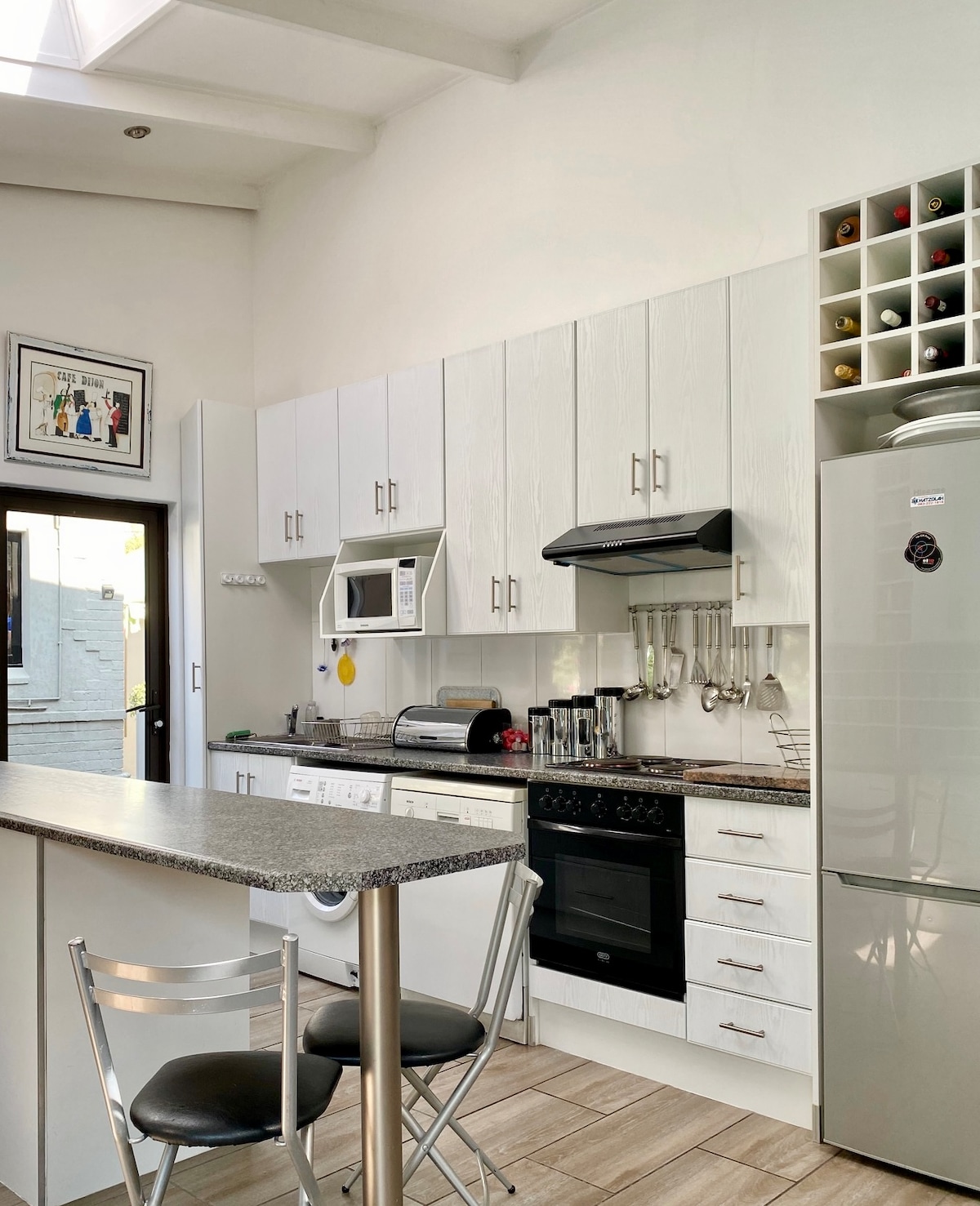 A modern kitchen features white cabinetry with ample storage, including a wine rack. Stainless steel appliances, including a microwave and oven, are present. A central island with seating for two provides additional workspace. Natural light enters through the glass door leading to the outside.