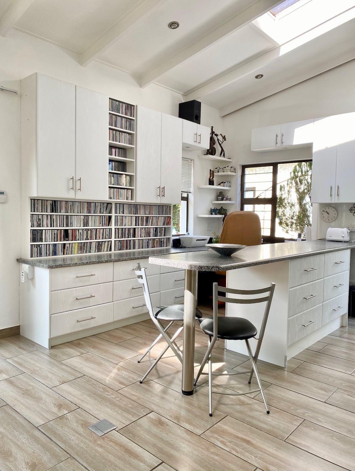 A bright kitchen space features white cabinetry and a sleek countertop. A compact dining table with two chairs sits near a large window, allowing natural light to fill the room. A collection of CDs is neatly arranged on a decorative shelf, adding character to the area.