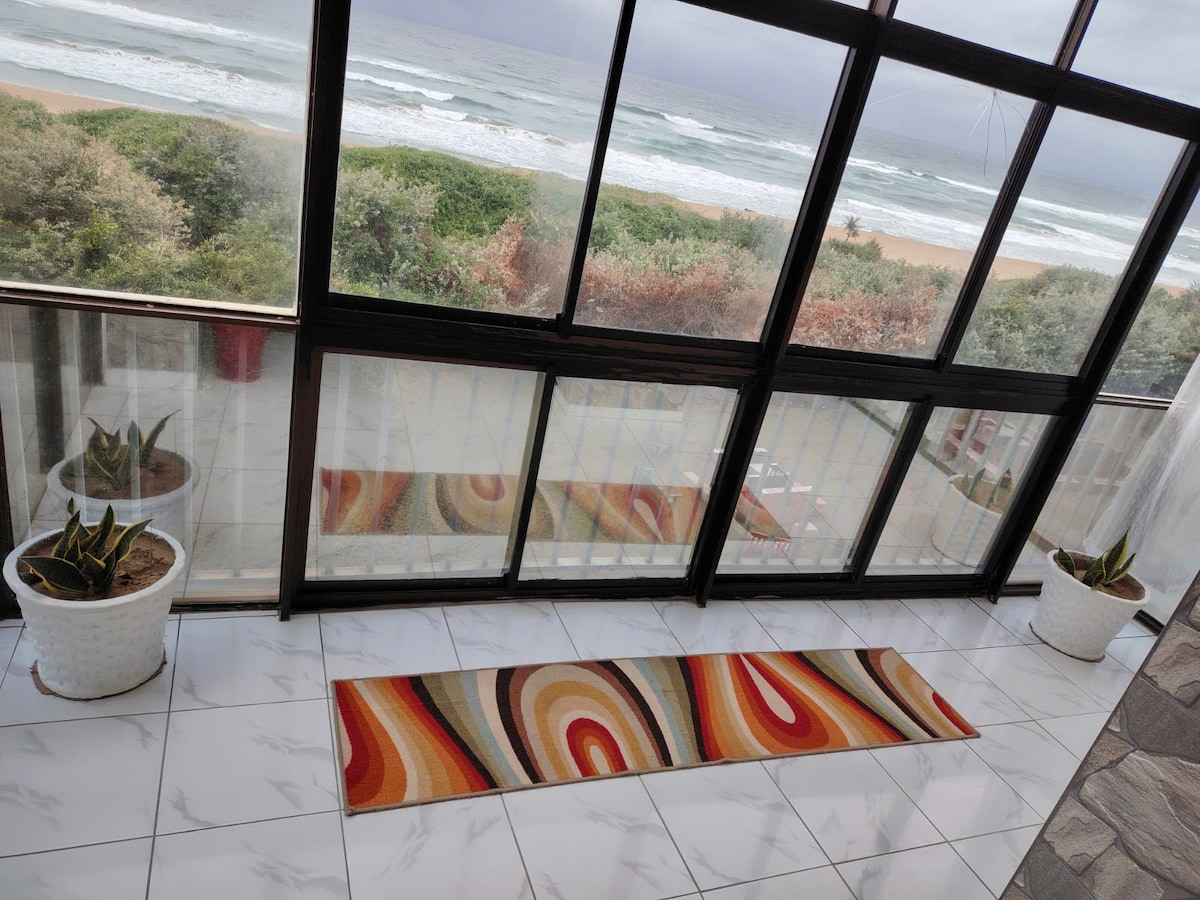 A bright sunroom features a colorful rug on white tiled flooring. Large glass windows provide uninterrupted views of the ocean and surrounding greenery. Potted plants on either side of the rug add natural elements to the space, enhancing the coastal atmosphere.