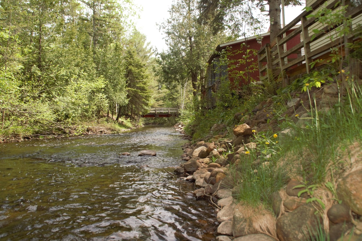 A serene creek flows gently alongside the cabin, bordered by smooth stones and lush greenery. Tall trees provide a natural canopy, while a wooden bridge is visible in the background, enhancing the tranquil setting.