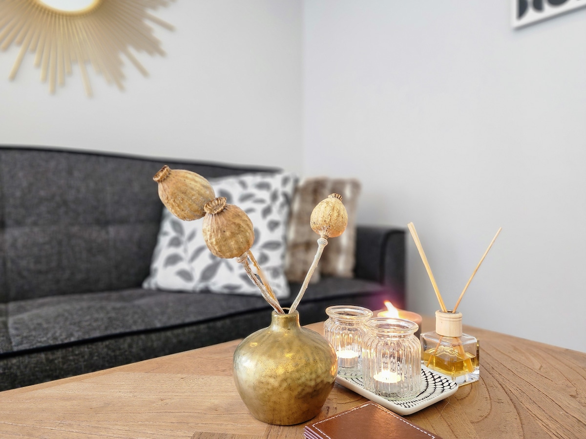 A decorative arrangement is displayed on a wooden coffee table, featuring a gold vase with dried plant stems alongside several glass candle holders emitting a warm glow. A stylish gray sofa with accent pillows is visible in the background, enhancing the inviting atmosphere.