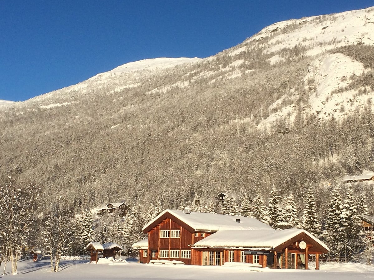 A large red wooden cabin is set against a backdrop of snow-covered mountains. The structure features multiple sections, large windows, and a welcoming entrance. Surrounding the cabin are tall evergreen trees and a blanket of fresh snow, creating a serene winter landscape.