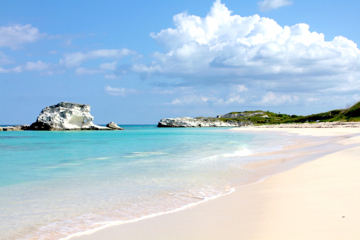 A sandy beach stretches along a tranquil coastline, with gentle waves lapping at the shore. Outcrops of white rock rise majestically from the clear turquoise water, under a sky filled with soft, white clouds, creating a serene coastal landscape.