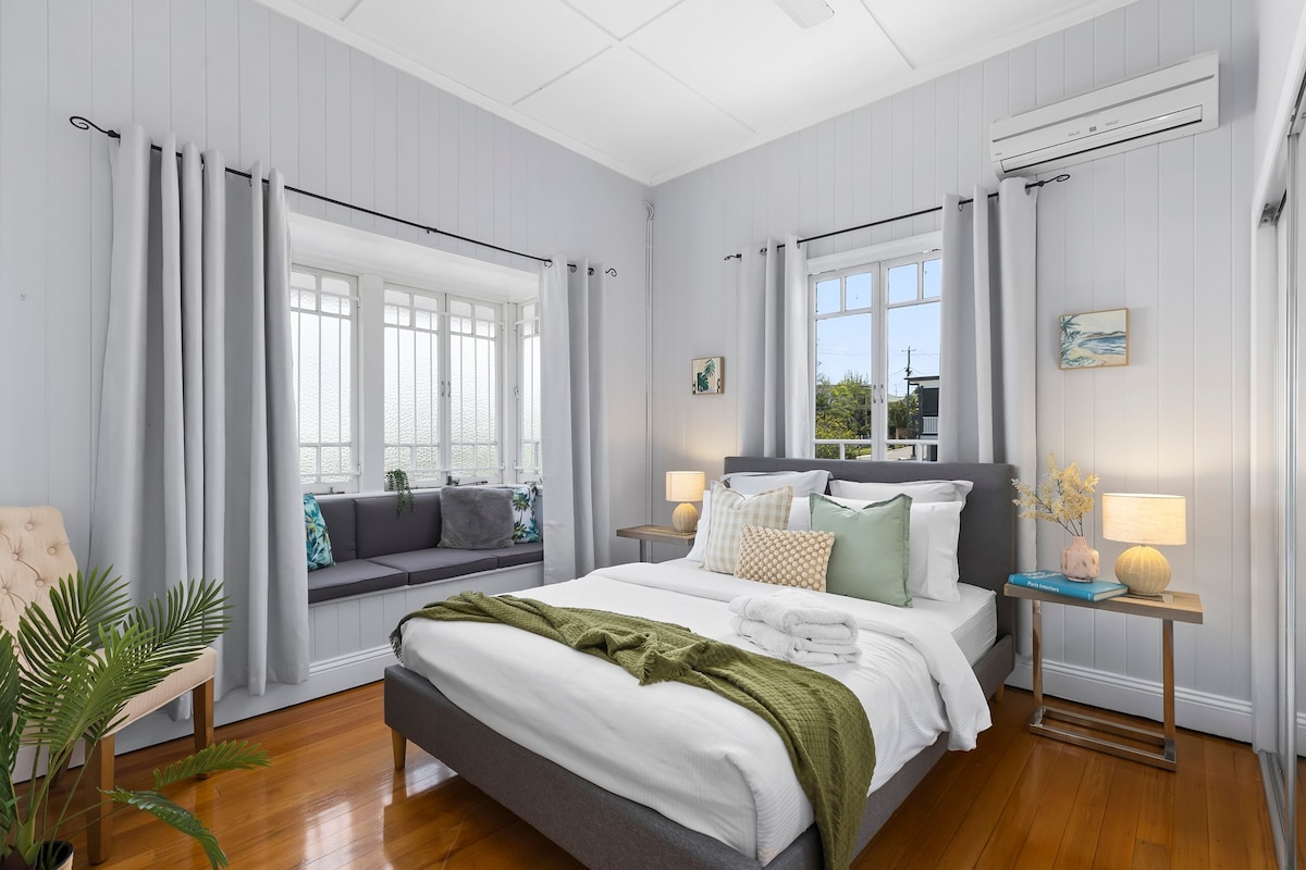 A well-lit bedroom features a spacious bed covered with soft linens and a textured throw. Natural light enters through large windows, complemented by elegant curtains. A cozy window seat provides additional seating, while bedside tables with lamps add functionality and a touch of warmth.