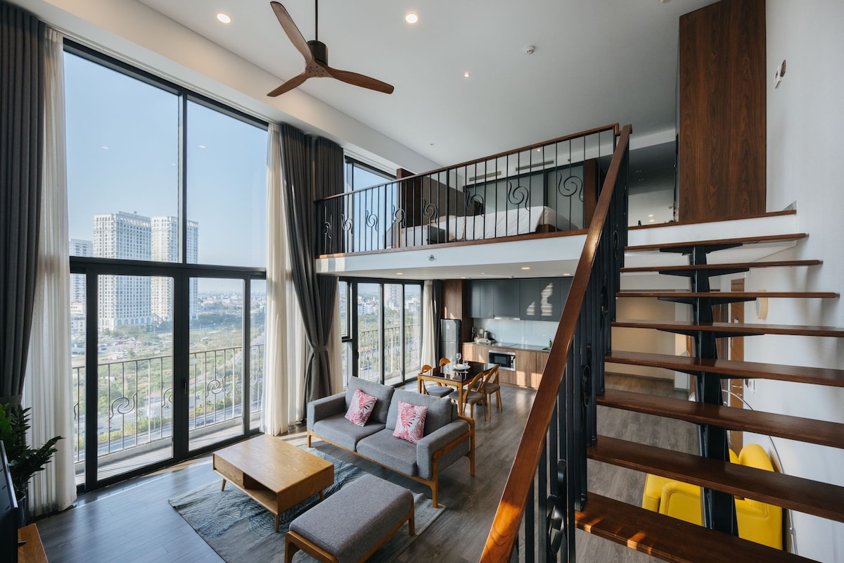 An open living area is seen with large glass windows offering city views. A wooden staircase leads to a lofted space. Comfortable seating is arranged around a wooden coffee table on a soft area rug, with plants adding natural elements to the décor.