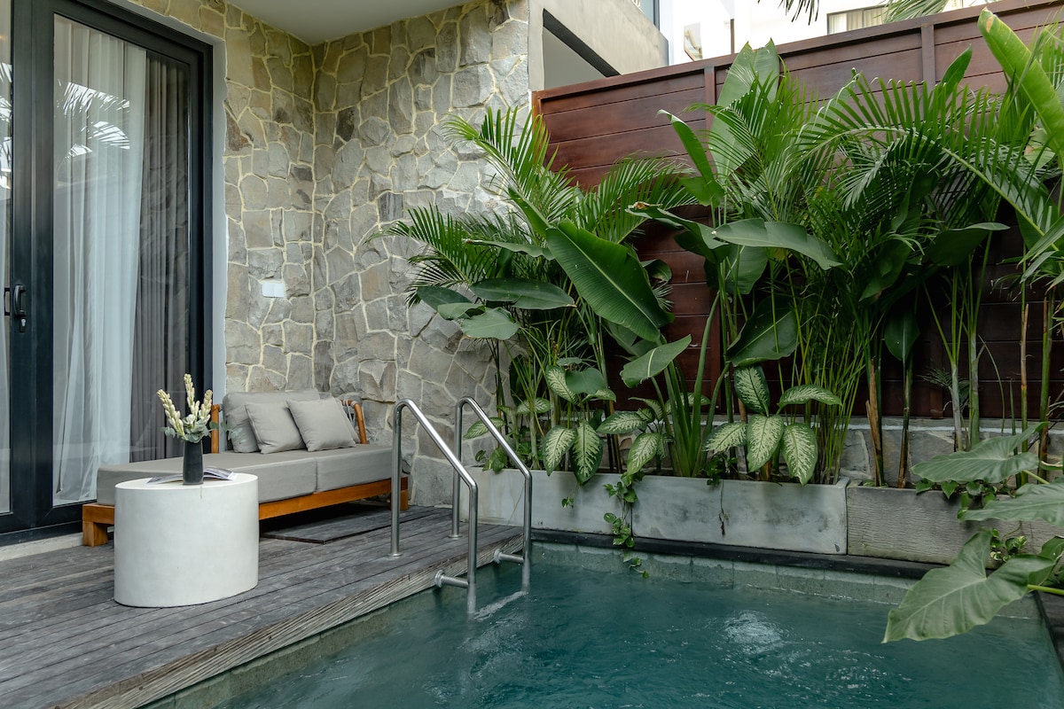 A private pool is surrounded by lush greenery, creating a serene atmosphere. A comfortable seating area is visible alongside the pool, featuring a cushioned sofa and a modern side table. The natural stone wall enhances the tranquil setting, inviting relaxation.