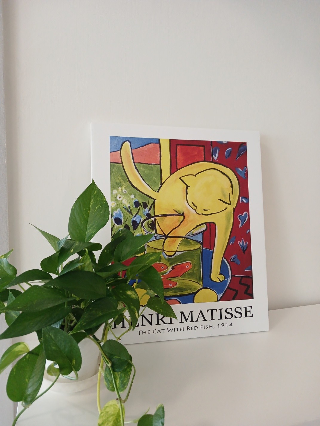 A colorful art piece by Henri Matisse titled 'The Cat with Red Fish' is displayed on a wall. Below it, a green plant adds a natural element to the decor, highlighting a blend of artistic and organic elements within the space.