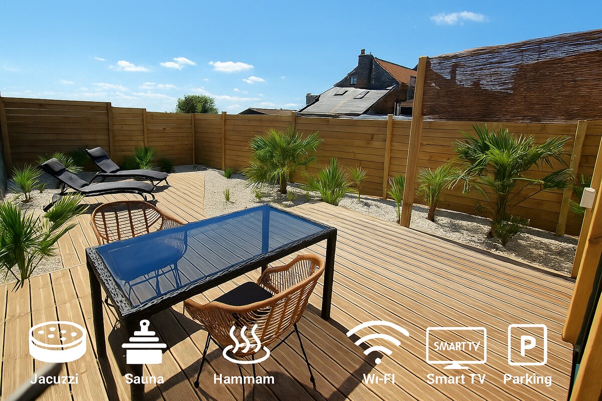 An outdoor terrace with wooden decking is surrounded by tropical plants. A glass-topped table is paired with woven chairs, and two lounge chairs are positioned nearby. The space is designed for relaxation, with a clear blue sky overhead and wooden fencing providing privacy.