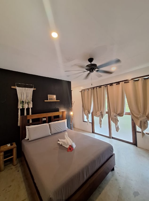 A cozy bedroom features a spacious bed with neutral-toned linens and decorative pillows. Large windows are adorned with light-colored curtains, allowing natural light to fill the room. A ceiling fan provides comfort, and a touch of greenery is present through the visible outdoor view.