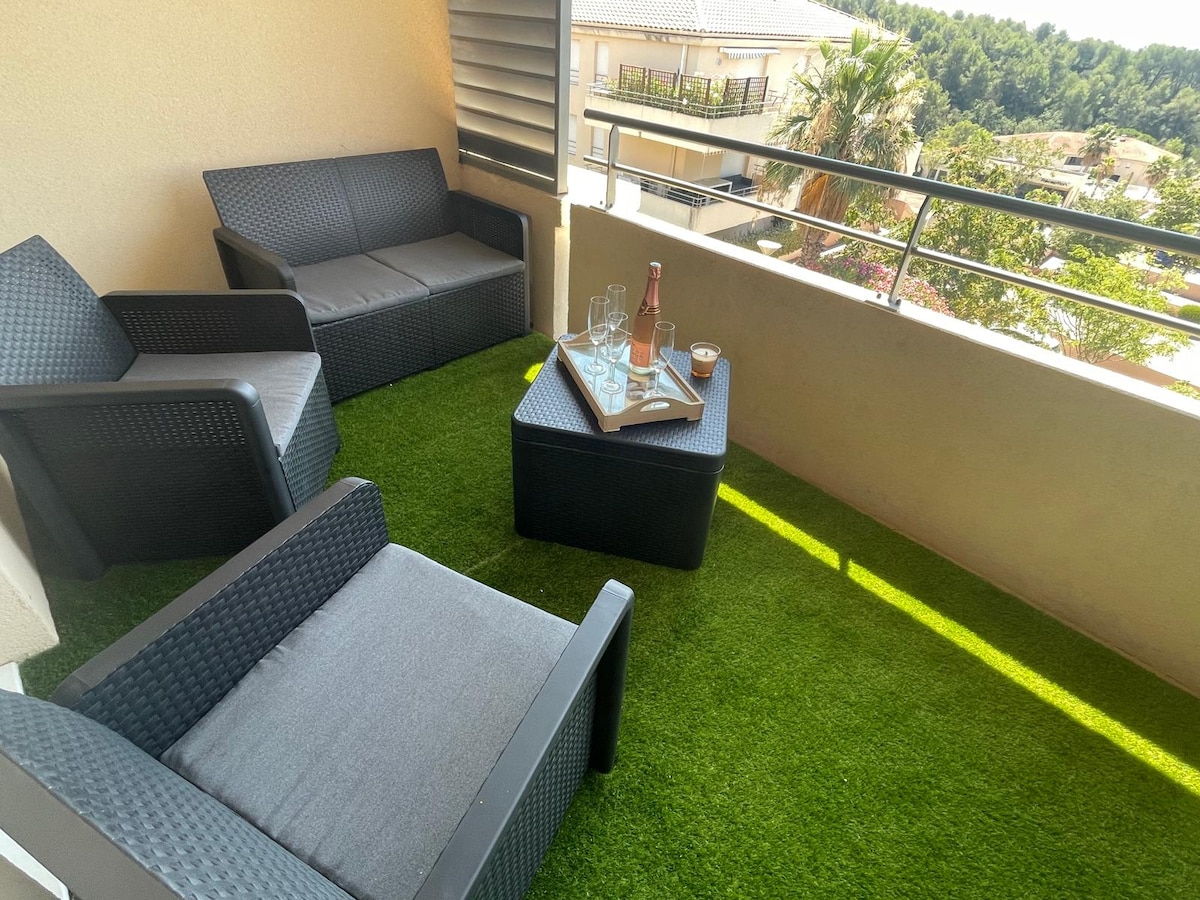 A cozy terrace features two seating areas with dark wicker furniture and gray cushions. A small table holds a bottle of rose wine and glasses. Lush green artificial grass provides a comfortable feel, while a view of surrounding trees can be seen in the background.