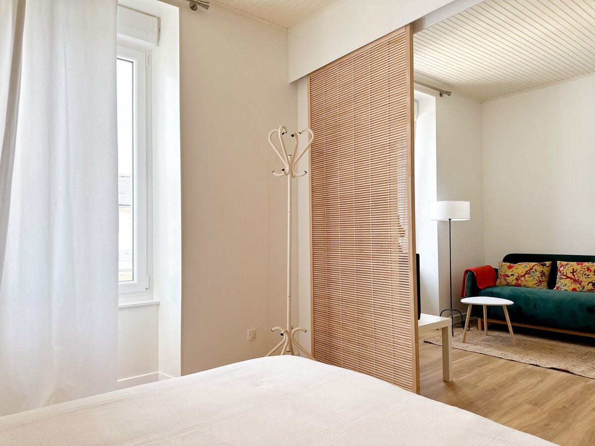 A bright bedroom area is visible, featuring a spacious bed with a textured white cover. A sliding partition, made of wood slats, separates the living area, which includes a colorful sofa and a small coffee table. Natural light enters through a window draped with sheer white curtains.