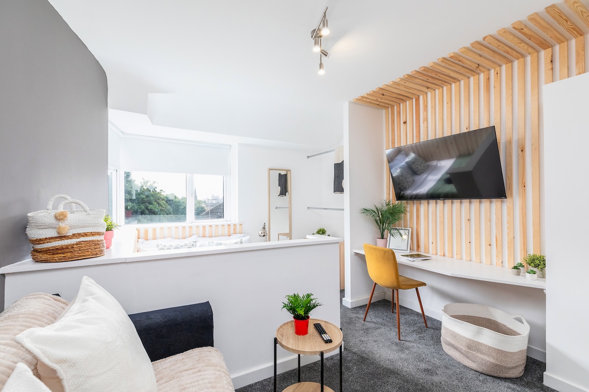 An open-plan living area is shown, featuring a cozy seating arrangement with a light-colored sofa and a small side table. A modern desk with a chair is positioned beneath a wall-mounted television. Natural light floods the space from large windows, complemented by a wooden accent wall.
