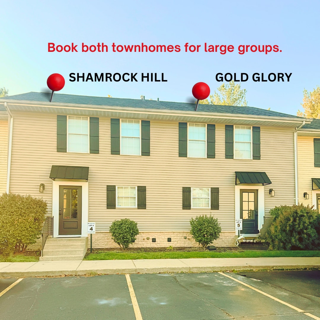 A view of the townhome exterior shows two units side by side with distinct signage. Each unit features dark green shutters and a covered entrance. A parking area is visible in front of the homes, framed by freshly paved asphalt.