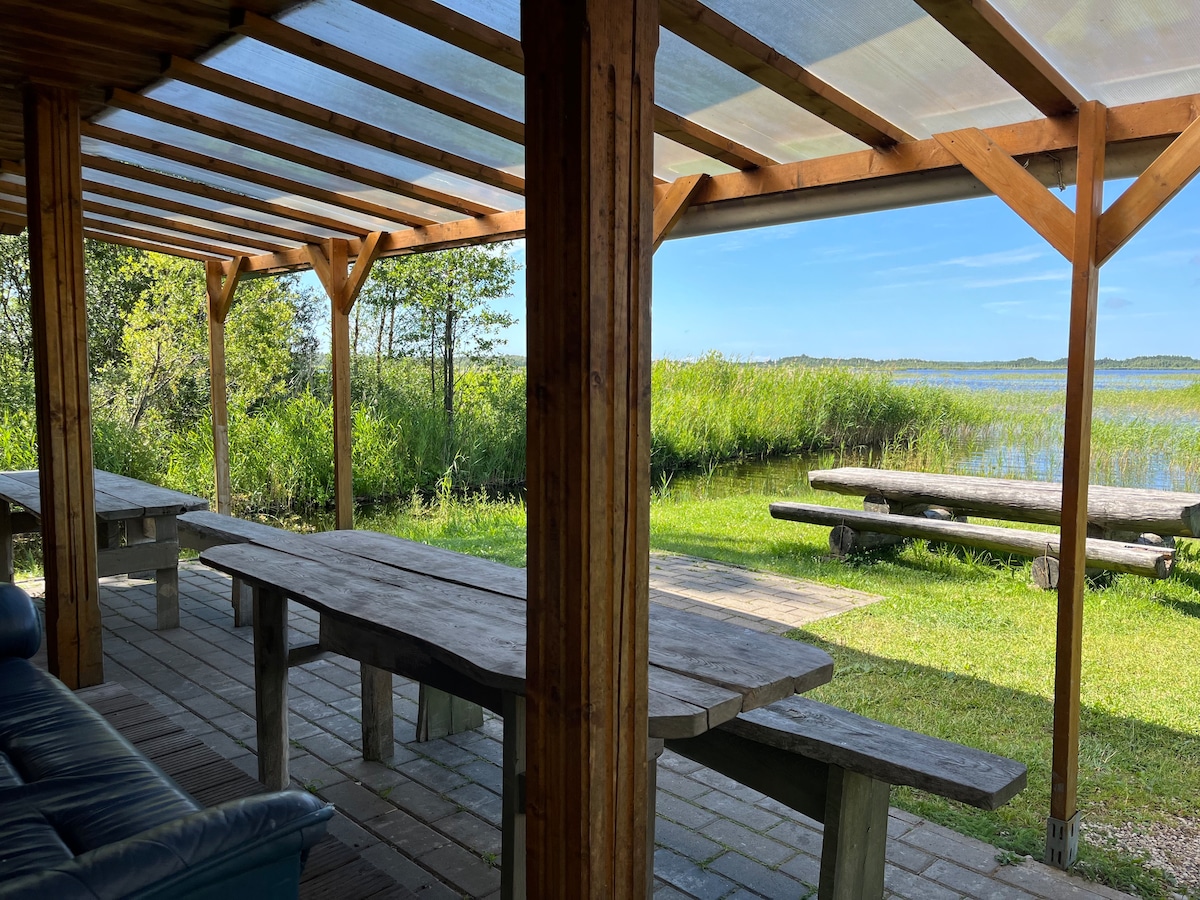 A covered patio area features sturdy wooden tables surrounded by greenery, with views of the lake beyond. Sunlight filters through the transparent roof, casting gentle light on the rustic seating. The tranquil natural setting is complemented by the sound of the water nearby.