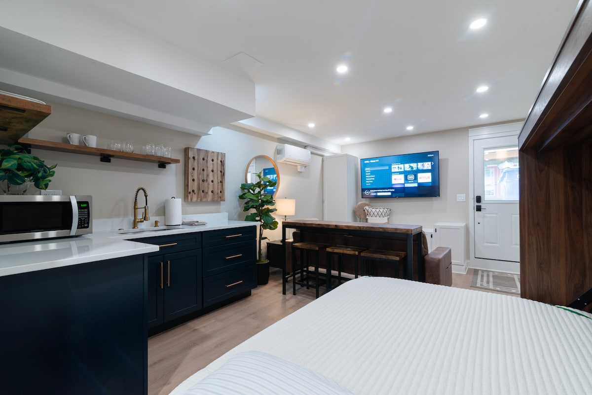 An open studio space is revealed, featuring modern cabinetry in navy with a white countertop. Comfortable seating surrounds a wooden table, and a large flat-screen TV is mounted on the wall. Soft lighting enhances the minimalist decor, and a door provides access to the exterior.