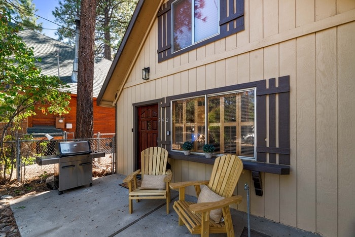 An inviting entryway is framed by two wooden chairs positioned on a concrete patio. A BBQ grill is situated nearby, and sunlight filters through the surrounding trees, enhancing the welcoming atmosphere of the outdoor space.