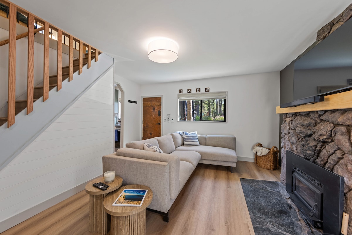 A spacious living area features a light-colored sectional sofa positioned near a stone fireplace. A circular ceiling light illuminates the room, with large windows providing views of the surrounding greenery. Decorative wooden tables sit nearby, adding a functional element to the cozy space.