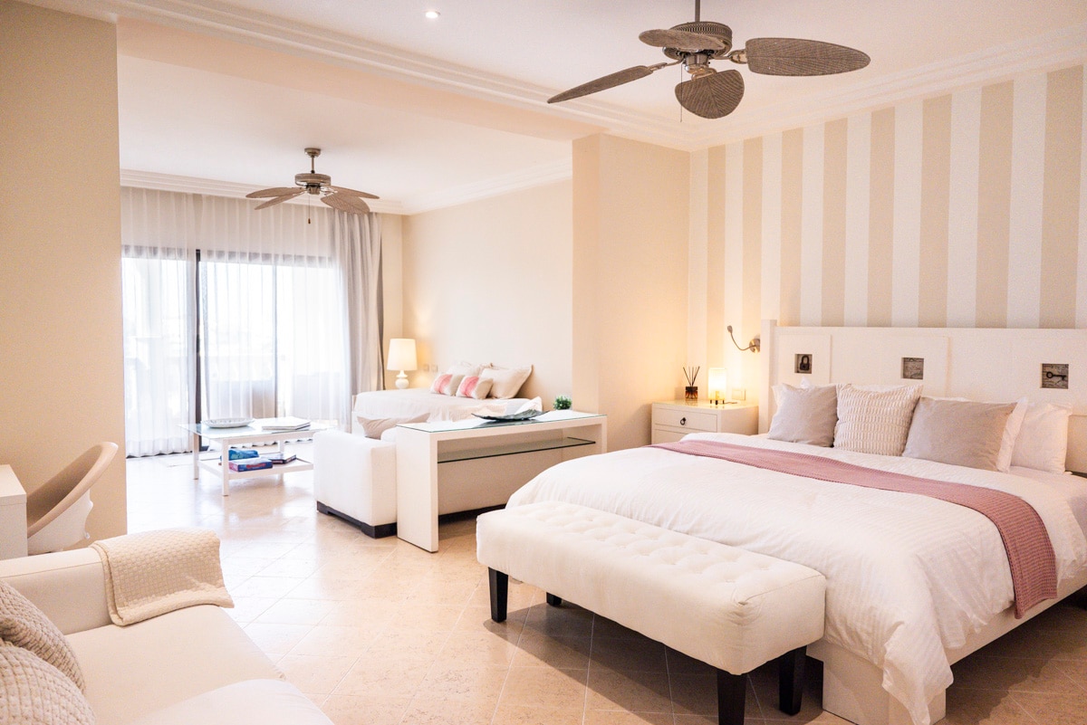 A spacious loft-style room is arranged with a comfortable king bed and a sofa area. Light colors and soft textures create a calming ambiance, while large windows provide natural light and a view of the outdoors. Ceiling fans add an additional touch of comfort.