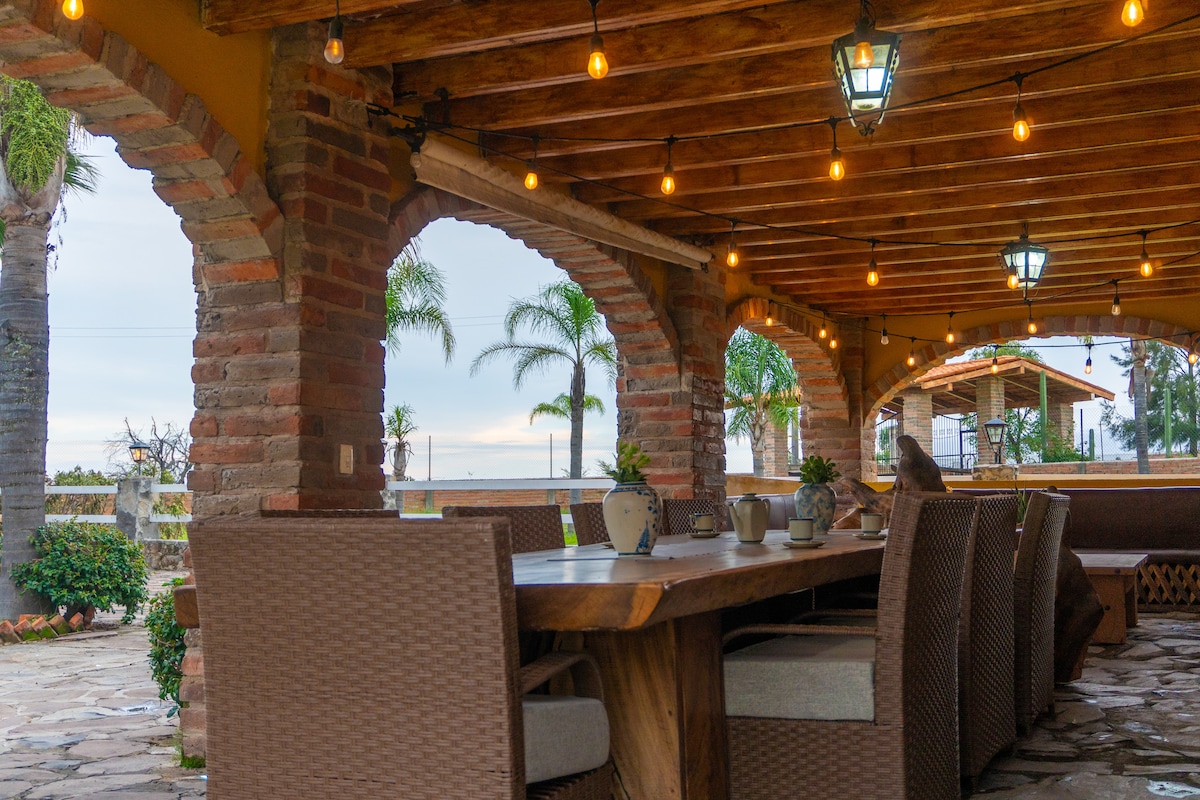 An outdoor dining area is framed by stone arches, featuring a long wooden table surrounded by woven chairs. Soft overhead lighting creates a warm ambiance, while palm trees are visible in the background, enhancing the serene atmosphere.