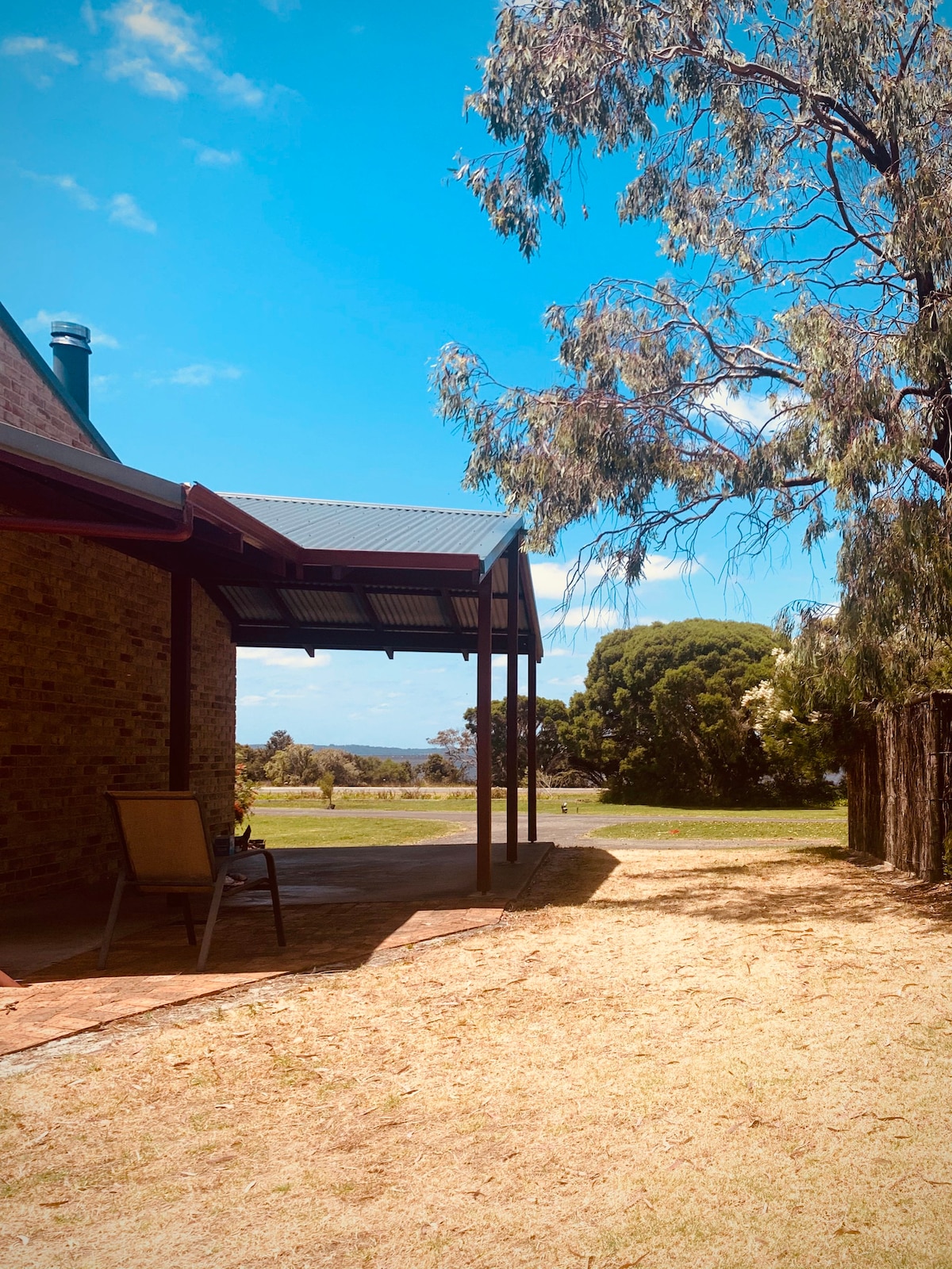 A private outdoor area is featured with a shaded patio, overlooking expansive lawns and gardens. A comfortable chair is positioned for relaxation. Lush greenery surrounds the space, and distant views of Wilsons Inlet can be glimpsed under a clear blue sky.
