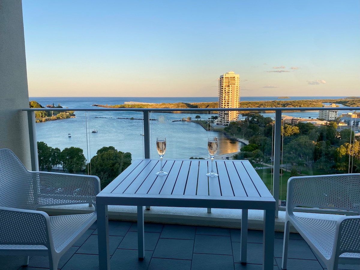 A spacious balcony is shown with a table and two chairs. Glasses of champagne sit on the table, facing scenic views of the ocean and the harbour. A distant building rises above the surrounding trees, completing the coastal landscape under a clear sky.