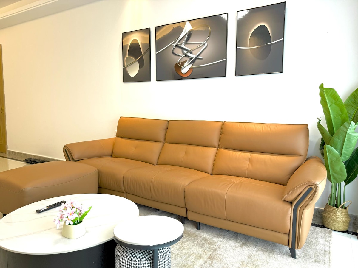 A spacious living area features a light brown leather couch with a modern design. Abstract artwork, framed in black, is displayed above the couch. A round coffee table with a floral centerpiece and a textured black and white side table are positioned in front, complemented by a large potted plant.