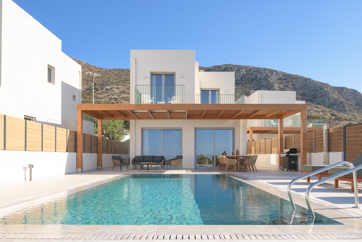 A modern villa features an expansive outdoor area with a shimmering pool. The wooden pergola provides shade over cozy seating and a dining area, all surrounded by a serene landscape. Large glass doors offer views into the villa's interior.