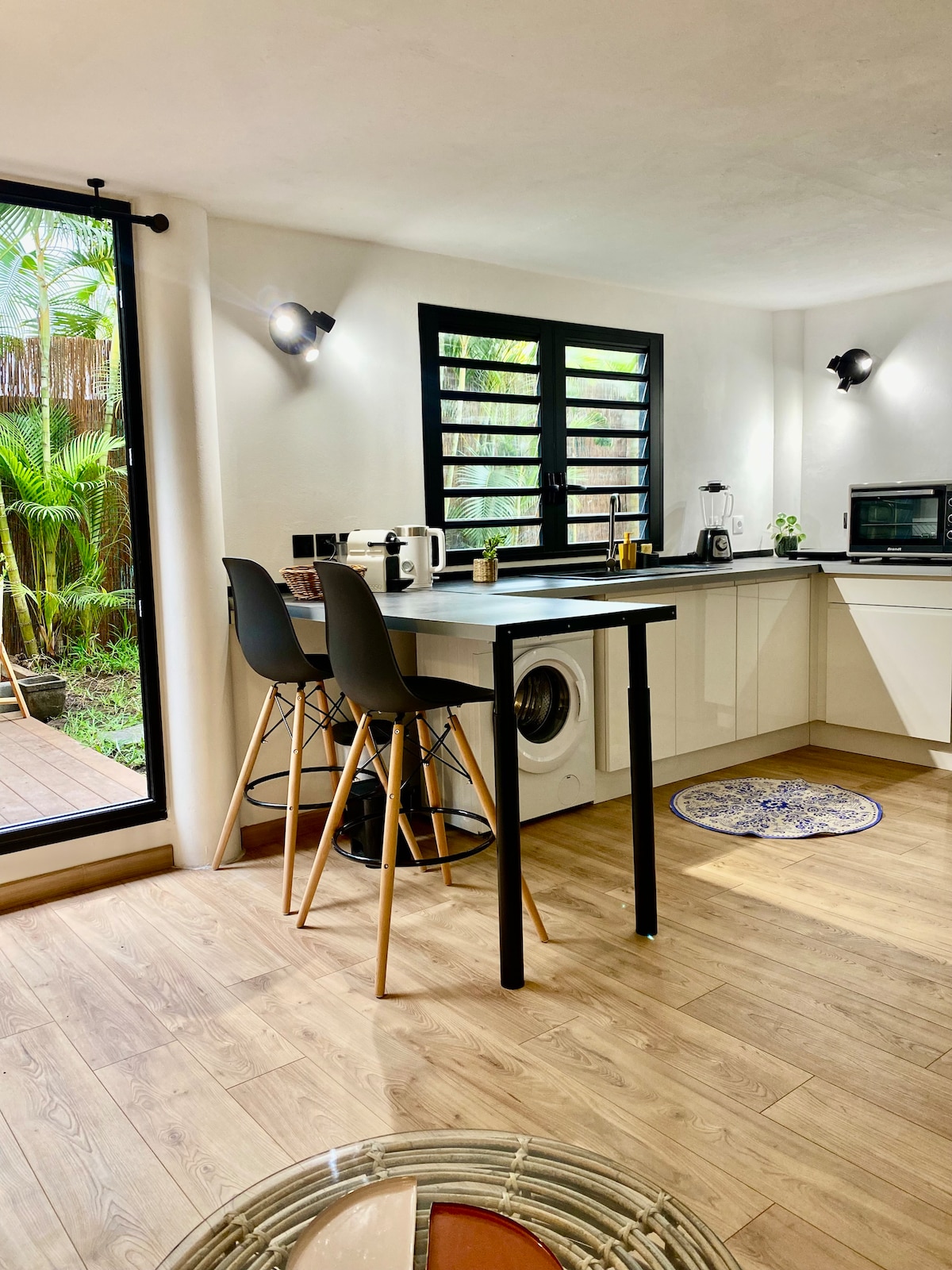 A modern kitchen area features a small dining table with two high-backed chairs. Natural light enters through a large window, showcasing greenery outside. The space includes essential appliances such as a microwave and a washing machine, positioned along clean, minimalist cabinetry.
