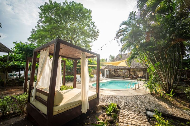A shaded relaxation area features a spacious daybed, draped with white curtains, surrounded by lush greenery. In the background, a swimming pool is visible, complemented by tropical palm trees and a seating area beneath a thatched roof.